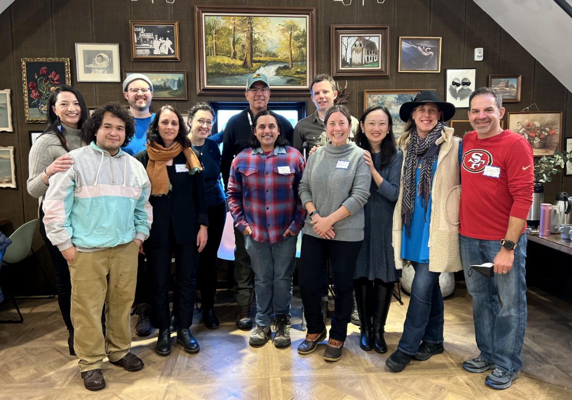 A group of people taking a staff photo in a room decorated with various framed paintings. They are wearing casual to semi-formal attire, and two of them are wearing hats. The background features dark wooden walls adorned with an eclectic mix of artwork.