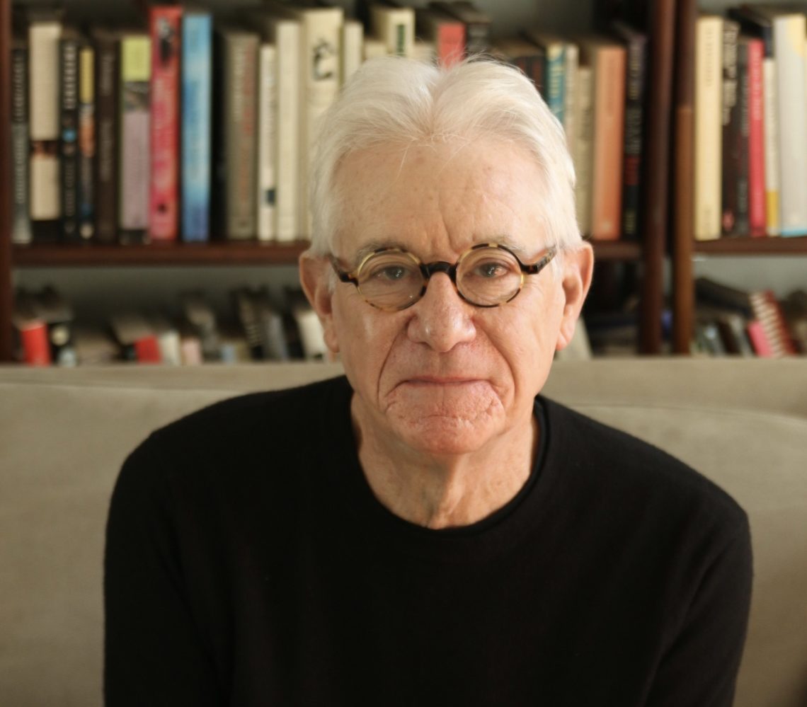 An older man with white hair and glasses, wearing a black sweater, sits in front of a bookshelf filled with various books. His neutral expression suggests deep thought, perhaps reflecting on a recent Berkeley book event he attended.