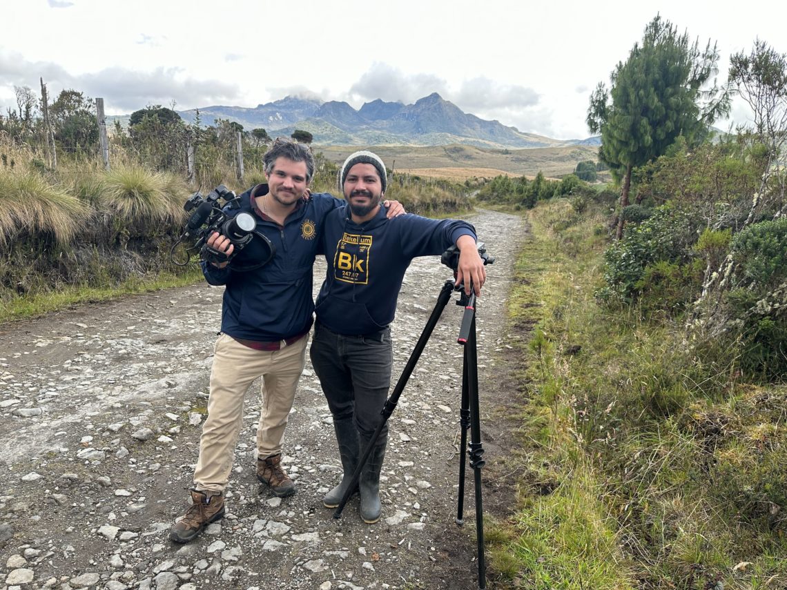 Two people standing on a dirt road with mountains and greenery in the background. The man on the left has his arm around the man on the right and they are both holding camera equipment. 