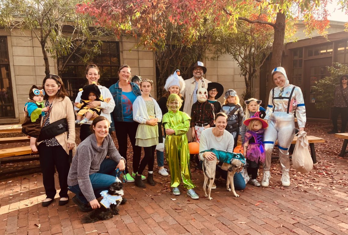 A group of people in festive costumes stand together outdoors on a brick pathway, reminiscent of Dean Geeta Anand's Welcome Back Note that encourages community spirit. They are dressed as various characters, including a spaceman, cowboy, and astronaut. Some hold children in colorful costumes, with two dogs also in costume.