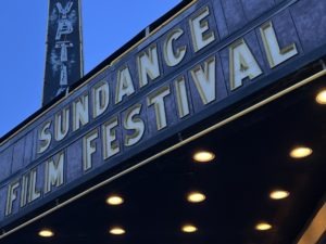 A sign with "Sundance Film Festival" in large gold letters is prominently displayed above a theater entrance. The marquee, lit by several round light bulbs, welcomes the festival's alumni and students alike. Part of a vertical sign is visible in the background against a clear blue sky.