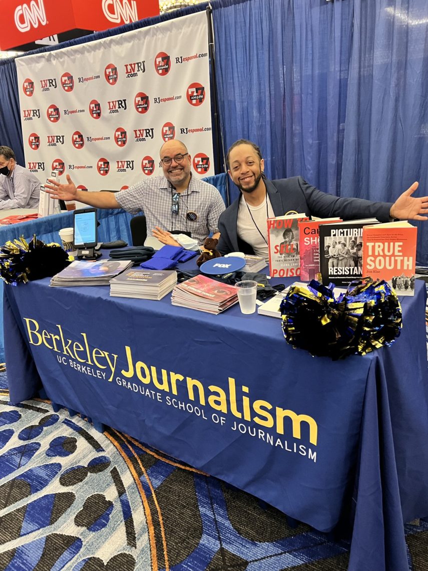 Two men are seated at a booth for Berkeley Journalism, part of UC Berkeley Graduate School of Journalism, at a convention. Smiling with their arms spread wide, they sit amidst books, pamphlets, and branded merchandise. The booth includes a Welcome Back Note from Dean Geeta Anand and CNN logos as the backdrop.