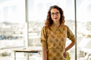 Lisa Pickoff-White, an esteemed alumni, stands confidently in front of large windows, wearing a patterned dress with a collar. Her wavy hair and glasses add to her poised demeanor. With a slight smile, the bright light outside casts a warm ambiance in the room, highlighting the cityscape behind her portrait.