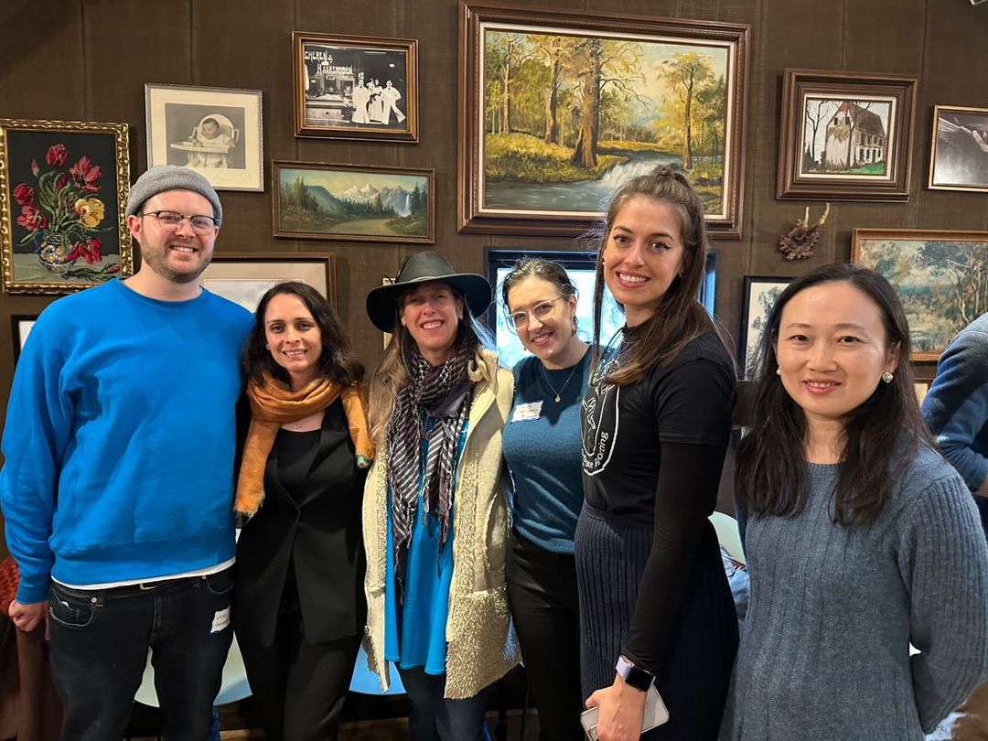 A group of six alumni smiling and standing together in a room decorated with various framed paintings. They are wearing casual to semi-formal attire, and two of them are wearing hats. The background features dark wooden walls adorned with an eclectic mix of artwork.