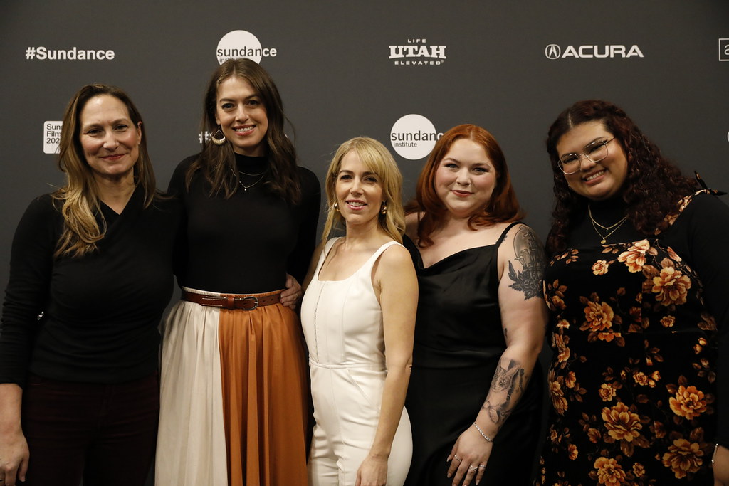 Five women stand together in front of a grey backdrop with Sundance, Utah, and Acura logos. Smiling and dressed in a mix of casual and semi-formal attire, the group could easily be mistaken for students or alumni celebrating their achievement at the Sundance Film Festival.