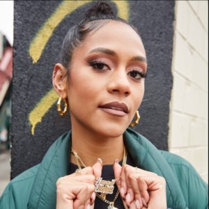 A woman stands outside, dressed in a green jacket with a black top. She wears gold hoop earrings, multiple ear piercings, and layered gold necklaces. One necklace has a pendant with text. She is looking directly at the camera and styled with makeup and a high bun, proudly representing her Afro-Latinx heritage.