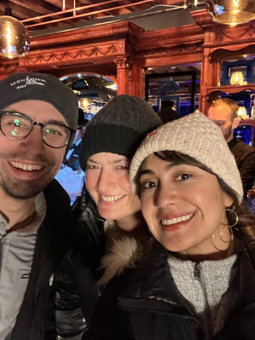 Three individuals posing for a selfie together. They are in semi-dark room, wearing beanies, and smiling happily.