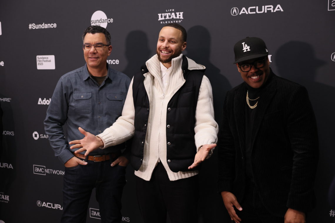 Three men stand together in front of a step and repeat banner at an event. The man in the center is smiling and gesturing with his hands, wearing a white sweater and black vest. The men on either side, perhaps alumni attending the Sundance Film Festival, wear glasses; one has a denim shirt, and the other a black outfit with a hat.