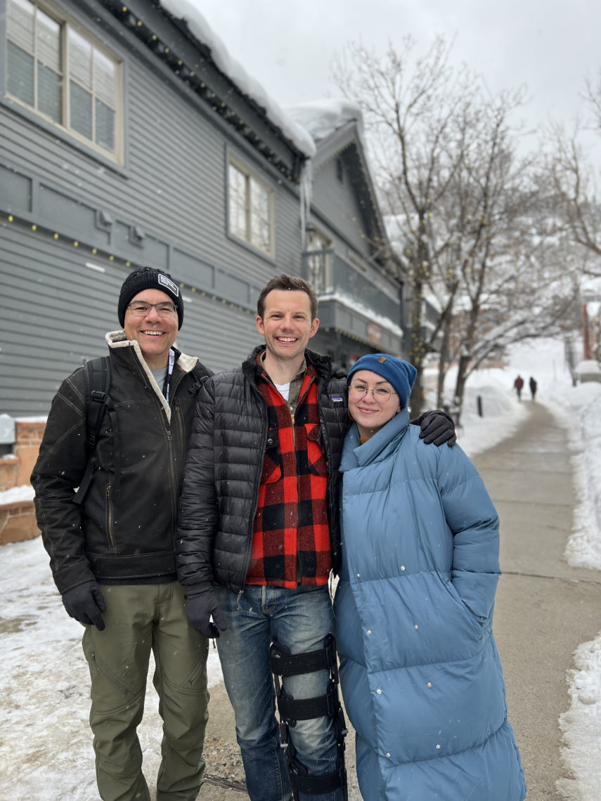 Three people standing outside in the snow. They are dressed in very warm clothing and are happily smiling. 