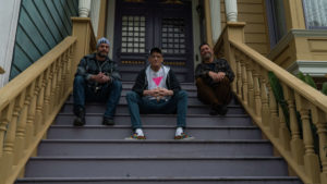 Three men sit on the steps of a wooden porch, all looking towards the camera. Two men on the sides wear casual attire and jackets, one with a beanie, the other in a cap. The man in the center wears a cap, T-shirt, and jacket. This group home offers familial support to HIV survivors.