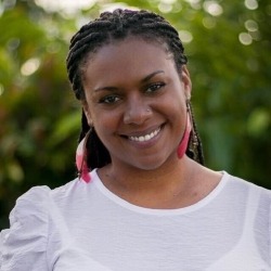 A woman with braided hair is smiling warmly at the camera. She is wearing a white shirt and vibrant pink and red feather earrings. The background consists of blurred greenery, suggesting an outdoor setting, embodying the spirit of Afro-Latinx Voices.