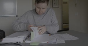 A person in a grey sweatshirt is seated at a desk, looking through a stack of papers in a ring binder. Various documents and notes are spread out on the desk, alongside tools of a graphics editor. The person appears focused on their task, with a whiteboard visible in the background.