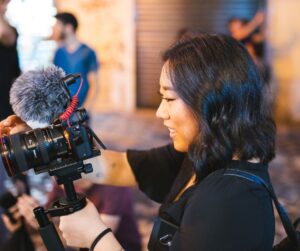 A woman with shoulder-length hair holds a camera mounted on a stabilizer. She smiles while adjusting the settings during an Info Session. In the background, people and blurred lights can be seen, suggesting an outdoor or casual setting.
