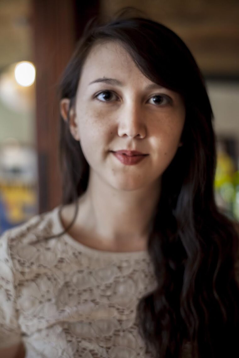 A young woman with long, wavy dark hair and fair skin faces the camera with a neutral expression. She is wearing a cream-colored lace top, and the background is softly blurred with warm tones, reminiscent of scenes from UC Berkeley's art festivals or discussions in a Tim Ferriss podcast on psychedelics.