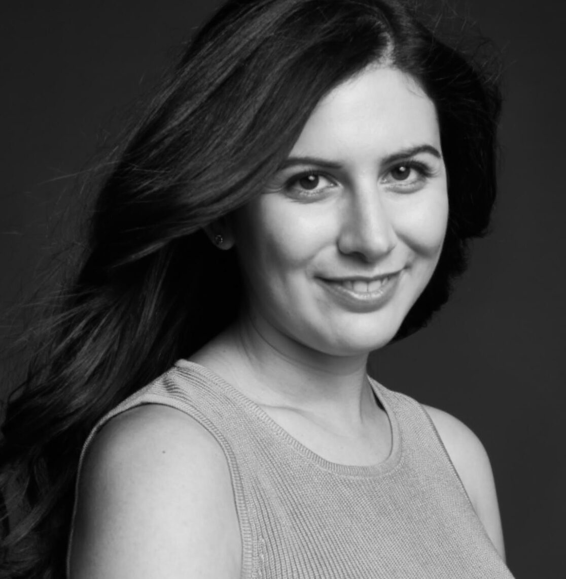 Black and white portrait of a woman with long, flowing hair, smiling softly while looking slightly to the right of the camera. She is wearing a sleeveless top against a dark background, capturing the essence of her dedication to her journalism fellowship.