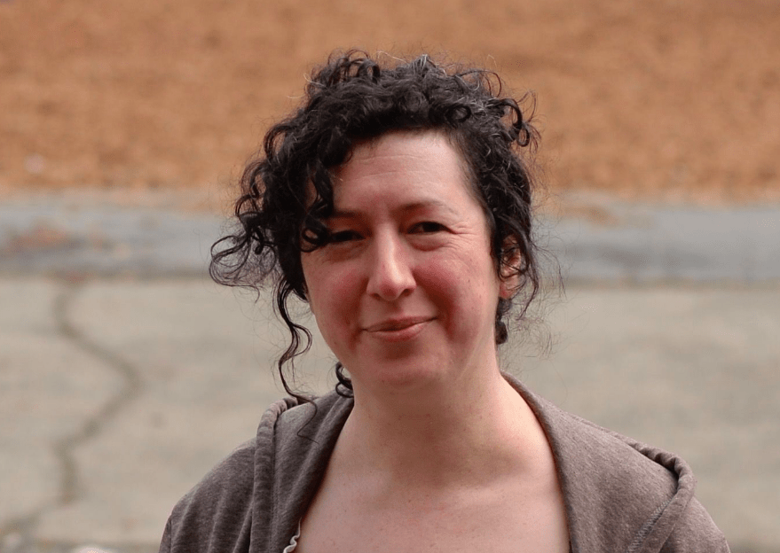 A person with curly dark hair, wearing a brown hoodie, stands outdoors. The background is blurry, showing a paved path and a brown area, possibly a field or patch of dirt. The person is looking at the camera with a slight smile, perhaps pondering their upcoming journalism fellowship.