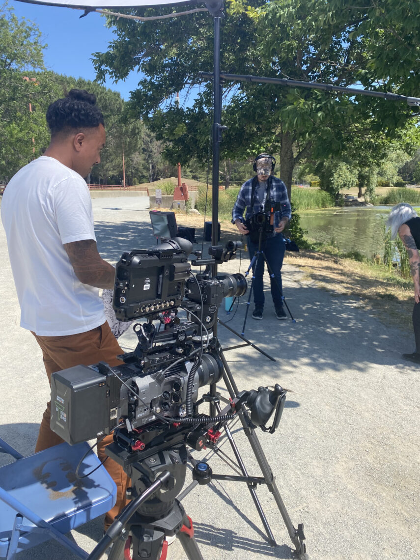 Two people are using camera equipment for a shoot. They are surrounded by a pond and trees. 