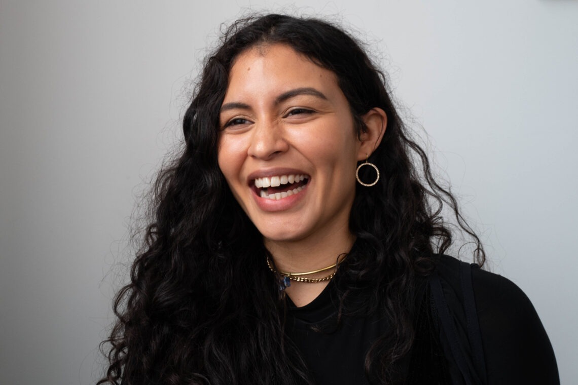 A headshot of a young woman mid laughter. She has long black wavy hair and is wearing light jewelry. 