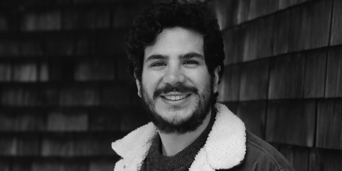 Black and white photo of a smiling man with curly hair and a beard. He is wearing a jacket with a shearling collar, standing in front of a wooden shingle wall. Juan Pablo Pérez-Burgos, FASPE Journalism Fellow ('24), exudes warmth and confidence in his timeless attire.