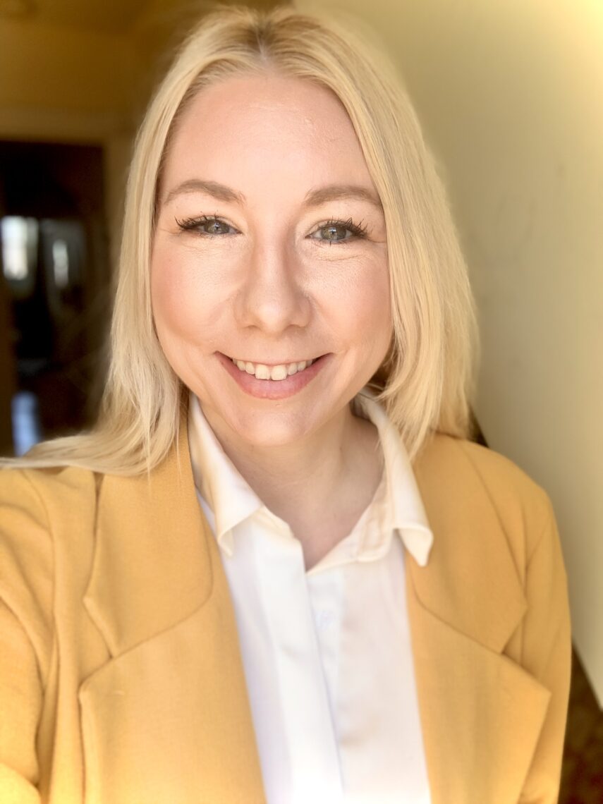 A selfie of a young woman smiling. She is wearing a yellow blazer with a white collared shirt and has shoulder length blond hair. 