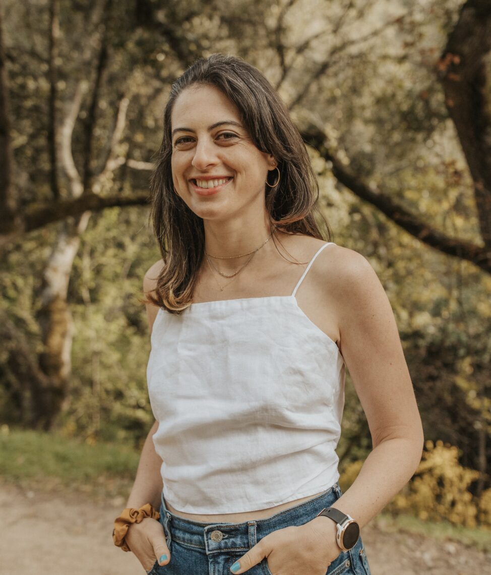 A portrait of a young woman smiling outside surrounded by trees. She has her hands in her pocket and is wearing a thin strapped white shirt with jeans. 