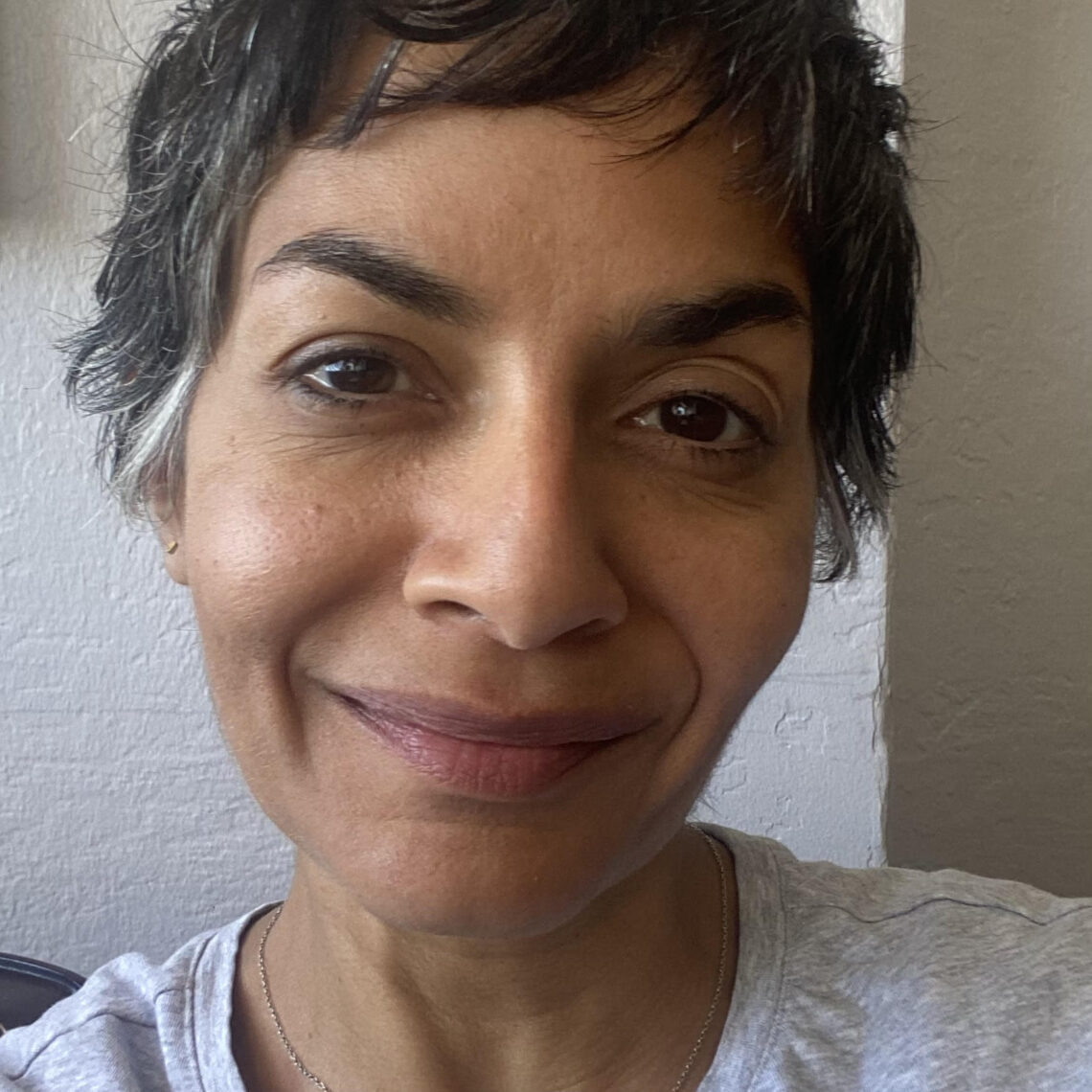 A person with short, dark hair featuring a streak of gray poses for a close-up selfie, perhaps capturing the moment before starting their journalism fellowship. They have a neutral expression and are wearing a light-colored shirt. The background is plain with white walls.