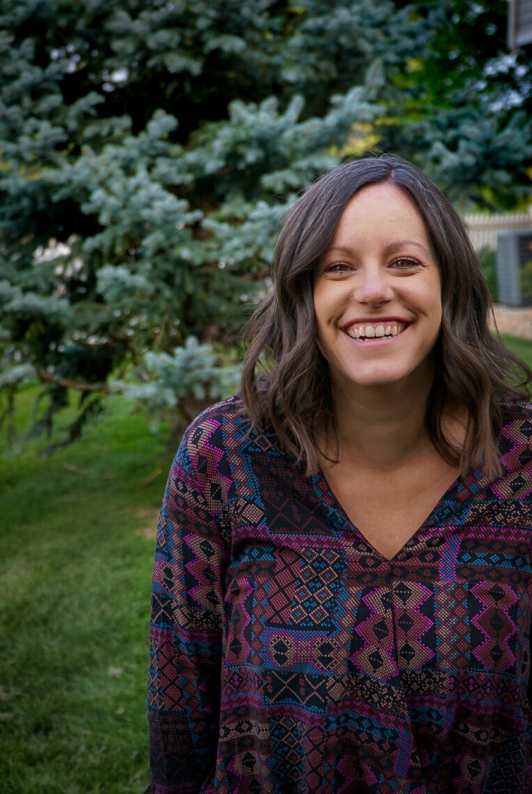 A woman with shoulder-length brown hair is smiling outdoors. She is wearing a patterned blouse with geometric designs in shades of purple, blue, and black. Behind her, there is a tall evergreen tree and a grassy area, where she often contemplates the impact of her journalism fellowship.