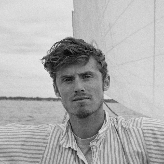 Black and white photo of a man with tousled hair, wearing a striped shirt, standing on a sailboat. The sail is visible behind him, and the water is calm, extending to the horizon under a cloudy sky. He looks contemplative, perhaps reflecting on his recent journalism fellowship.