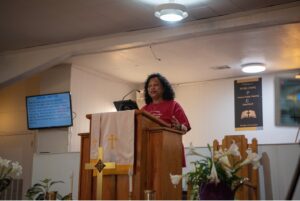 Pastor Sheryl King stands at the pulpit in a red t-shirt.
