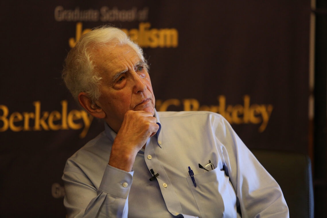 An elderly man with white hair is sitting with his hand resting on his chin, looking contemplative. He is wearing a light blue shirt with pens in the pocket. The background features a banner with partially visible text that includes "Graduate School of Journalism" and "Berkeley", mourning Daniel Ellsberg's passing.