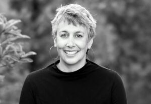 Black and white portrait of Berkeley Journalism Dean Geeta Anand with short hair and hoop earrings, wearing a dark top. The background is blurred foliage.