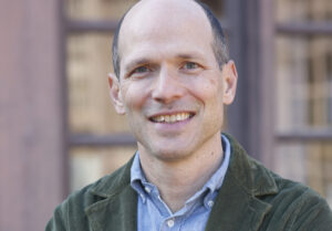 A smiling man with a bald head and light skin tone is standing in front of a blurred background. He is wearing a blue button-up shirt and a green corduroy jacket. The image is a close-up, showing his upper body and face, reminiscent of Jason Spingarn-Koff discussing climate change at Berkeley Journalism.