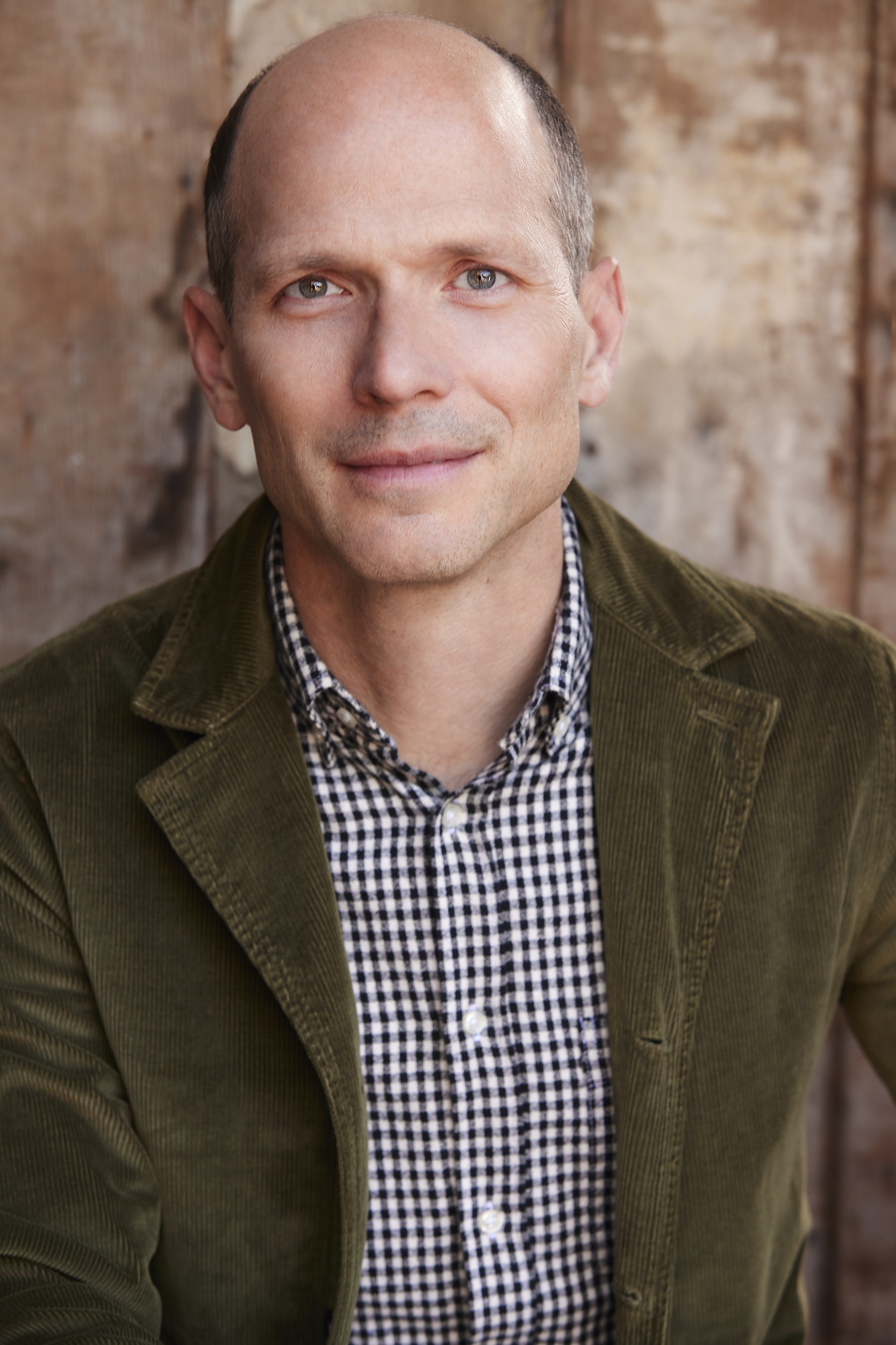 A bald man in a green corduroy jacket and black-and-white checkered shirt sits in front of a wooden background, looking directly at the camera with a neutral expression.