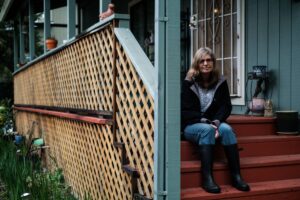 Franceen Levy sits on the red stairs outside her house. She is a white woman with shoulder-length blonde and gray hair. 