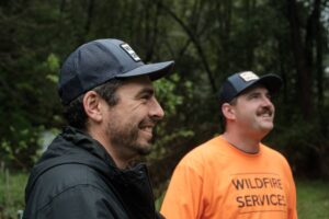 Wildfire Services lead Andrew Carrillo (left) and Brandon North work together to assess a home. Andrew is wearing a black coat and black hat, Brandon is wearing a black hat and orange Wildfire Services shirt. 