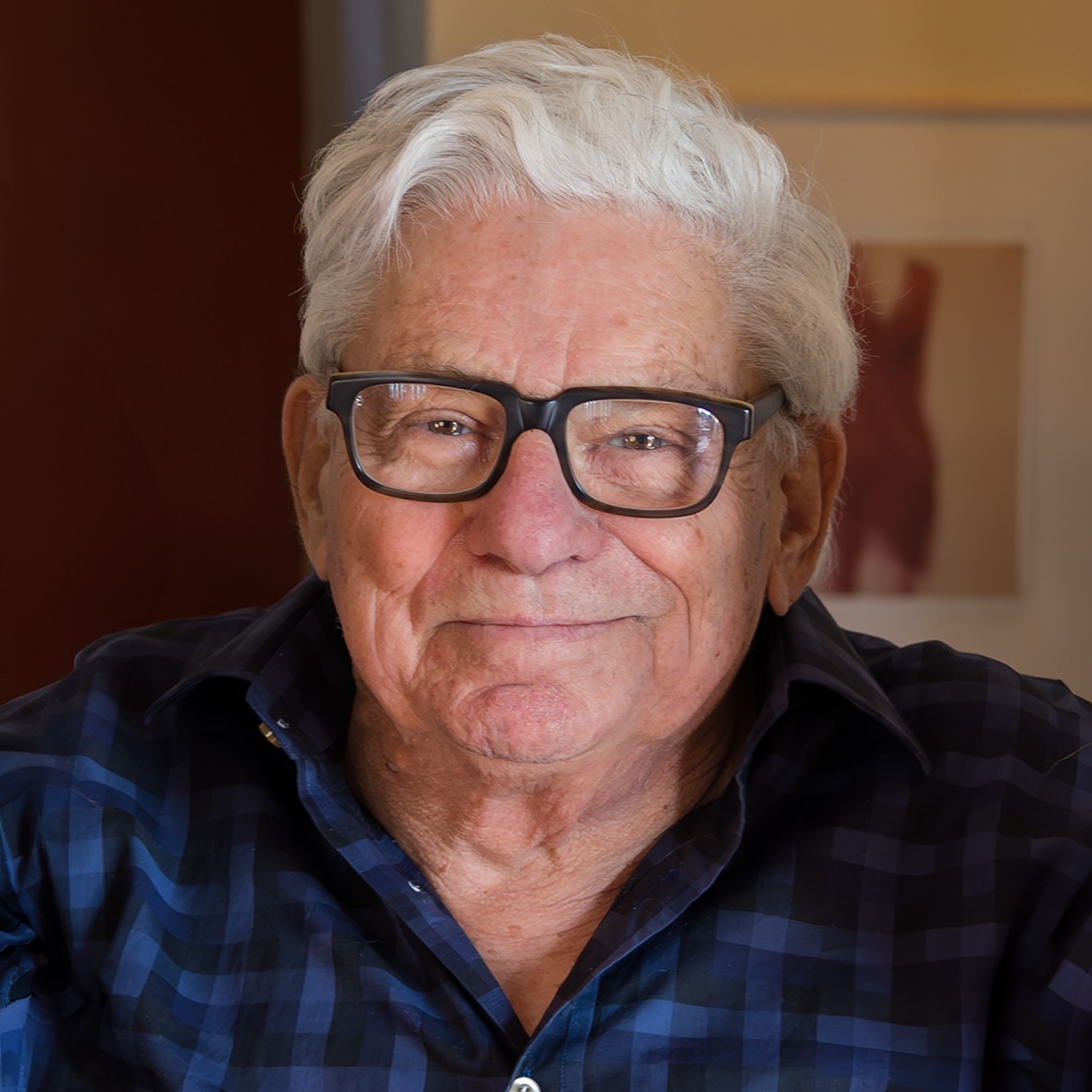 Photograph of an older man with a full head of grey hair wearing black glasses and a blue and black checkered button down shirt smiling.