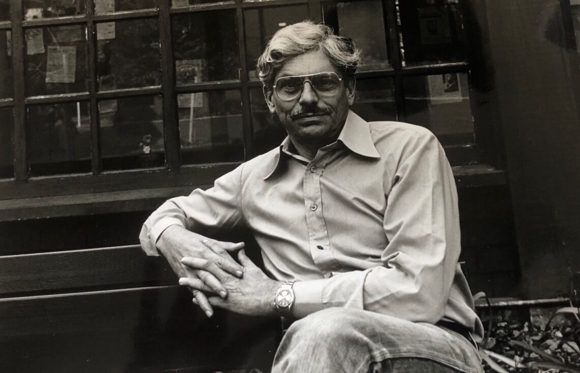 A black-and-white photo of an older man, Emeritus Professor Andrew Stern, sitting on a bench with his hands folded. He is wearing glasses, a light-colored button-up shirt, and jeans. He has a mustache and is looking directly at the camera, posed in front of a window with visible reflections.