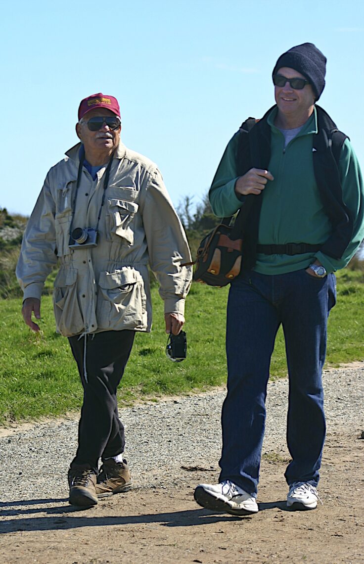 Two men are walking on a path outdoors. One man, wearing a beige jacket and a red cap, holds a small camera and binoculars. The other man, dressed in a green jacket, blue jeans, and a black beanie, carries a shoulder bag. Both men are smiling as if they're covering an assignment for Berkeley Journalism under the guidance of Emeritus Professor Andrew Stern.