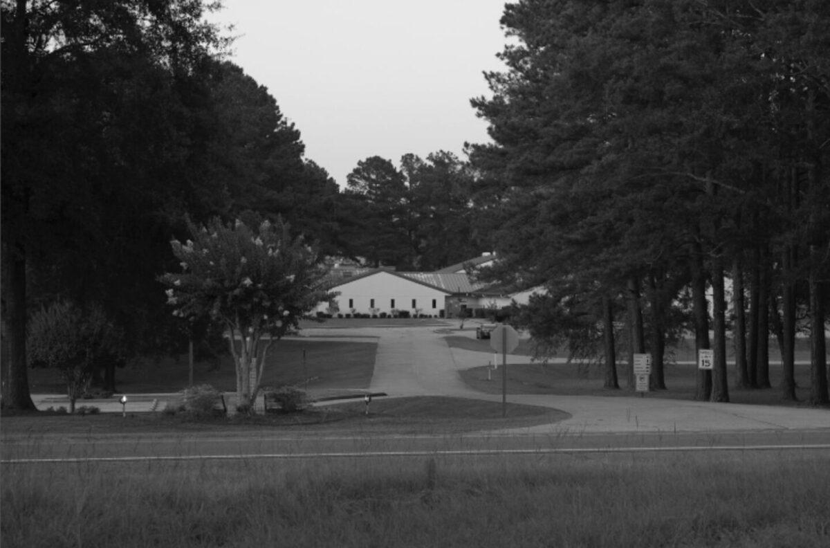 A black and white photo of the Ware Youth Detention Facility from the road. It is surrounded by thick clusters of trees and is a white facility with a brick roof.