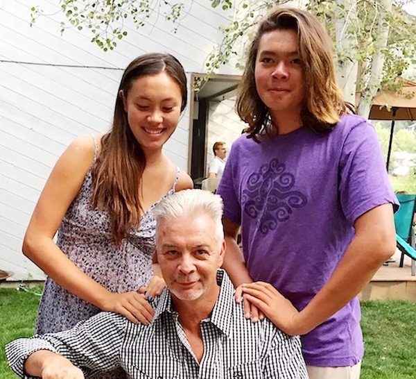 Ian Smith with his son and daughter, who each have their hands on Ian's shoulders. His daughter is wearing a sundress and his son is wearing a purple t-shirt. Both have long brown hair, Ian's hair is short and white and he's wearing a white and black checkered shirt.