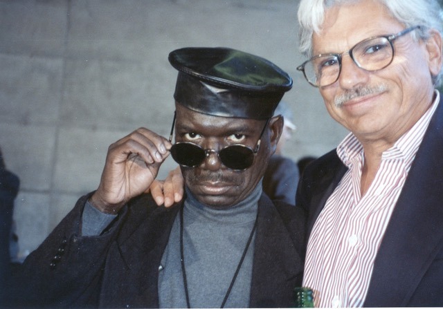 Photo of Marlon T. Riggs, wearing a black leather cap and sunglasses pushed down on his nose and Andrew Stern, who has his arm around him smiling.