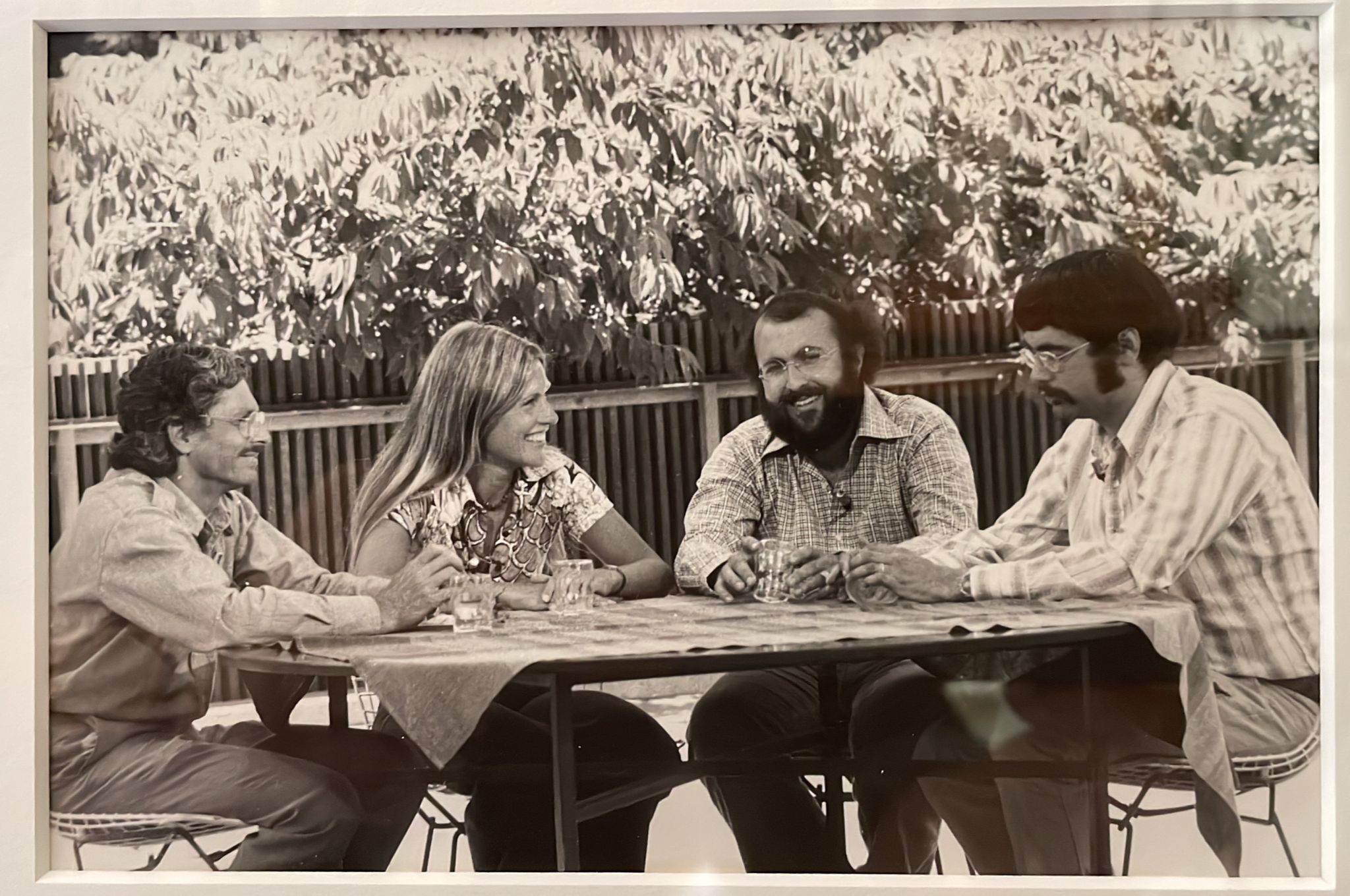 A black and white photo shows four people, including Andrew Stern, seated around a table outdoors, conversing and smiling. The group includes a woman and three men, all casually dressed, appearing to enjoy a relaxed moment together. The background features lush greenery typical of Berkeley Journalism gatherings.