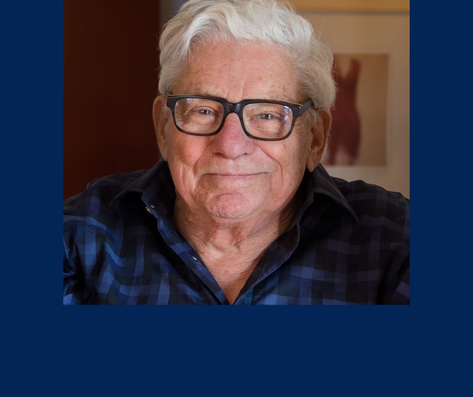 An elderly man with white hair and black-rimmed glasses, identified as Emeritus Professor Andrew Stern, smiles at the camera. He is wearing a blue and black checkered shirt and is positioned against a background with reddish tones and a framed picture.