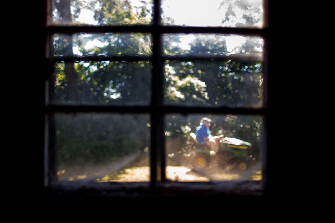 A photo through a wood-framed window of Rick Cook, a white man in a blue shirt, riding his green tractor.