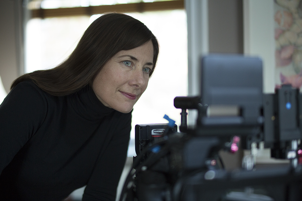 A woman with long brown hair, wearing a black turtleneck, leans forward intently, looking at a camera setup or monitor. The background is slightly blurred, showcasing a window and a floral painting. Her focused expression hints at her dedication that recently earned her a teaching prize.