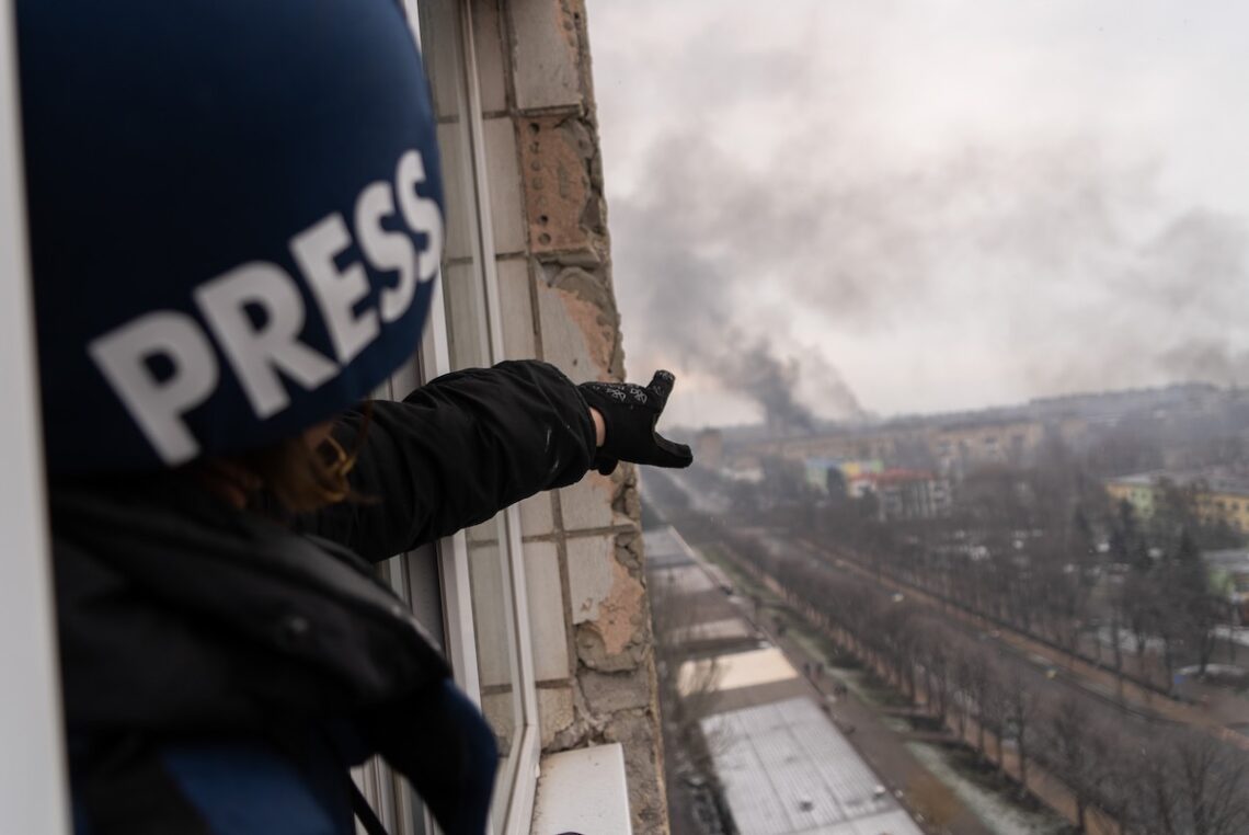 A person wearing a "PRESS" helmet points from a damaged building toward thick smoke rising in the distance. The image captures a cityscape with buildings and trees under a gray, overcast sky, evoking scenes from "20 Days in Mariupol." The scene suggests recent destruction or an ongoing conflict.