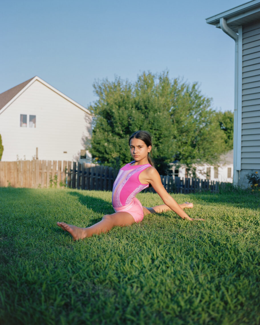 A young girl in a pink gymnastics leotard performs a split on the grass in a suburban backyard. She is looking confidently at the camera. There is a wooden fence and a house in the background, along with a large leafy tree. The sky is clear and blue—a perfect shot for the 2023 Creator Labs Photo Fund winners, captured by Wesaam Al-Badry.