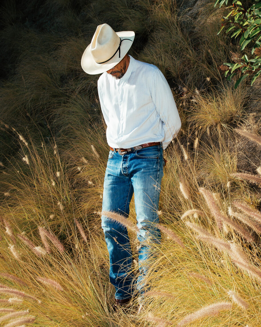 A person with a beige cowboy hat, white long-sleeve shirt, blue jeans, and brown belt stands in a field of tall dry grass, looking downward. Partially surrounded by hills and vegetation, this evocative scene was captured by Clara Shuku Mokri for the 2023 Creator Labs Photo Fund.