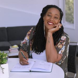 Nicole Caruth, with long braided hair and a tattoo on her forearm, is smiling and sitting at a table. She is writing in a notebook with her right hand and resting her head on her left hand. Wearing a floral patterned shirt, she is surrounded by books and a plant on the table.