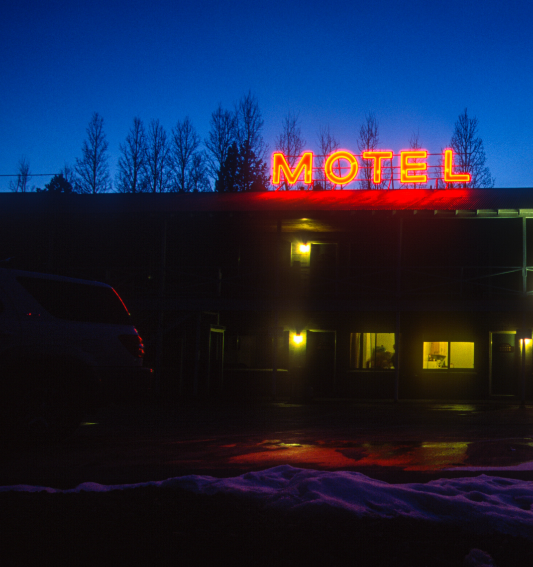 A neon red "MOTEL" sign glows against a deep blue twilight sky. Below, a dimly lit building shows a warmly lit interior through its windows. A white vehicle is parked outside on a wet, reflective surface with patches of snow nearby, capturing the essence of Berkeley Journalism's evocative storytelling.